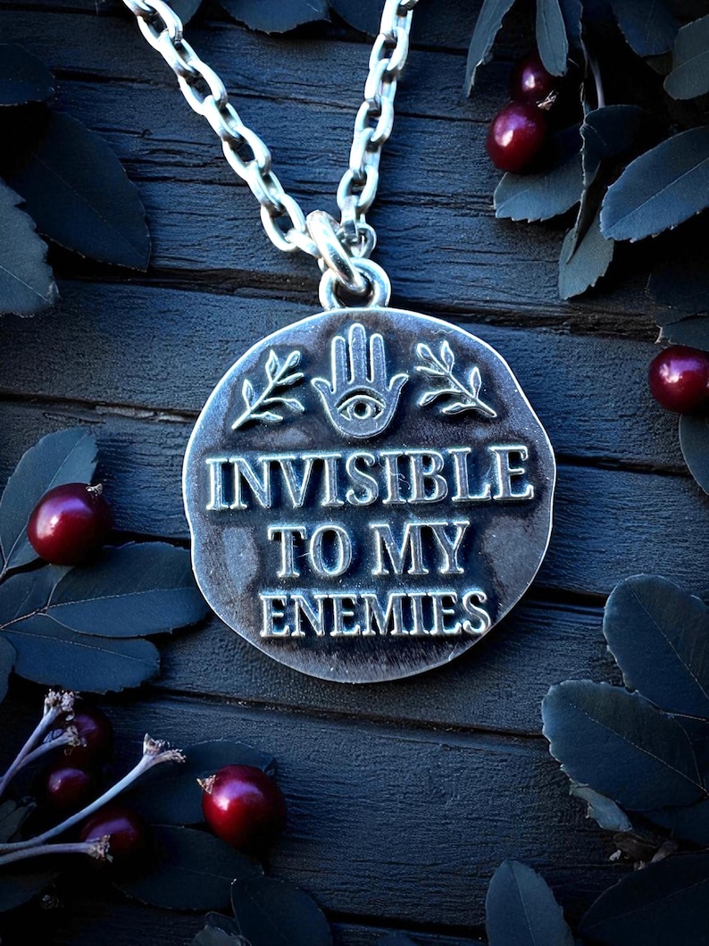 Sterling silver pendant engraved with the phrase ‘Invisible to My Enemies,’ featuring an all-seeing eye in a hand and leafy branches, shown on dark wood with berries and leaves