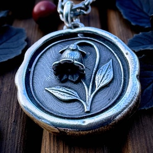 May include: A silver-toned pendant necklace featuring a floral design. The pendant has a circular shape with a raised border and a detailed image of a flower with leaves. The chain is also silver and has a link design.