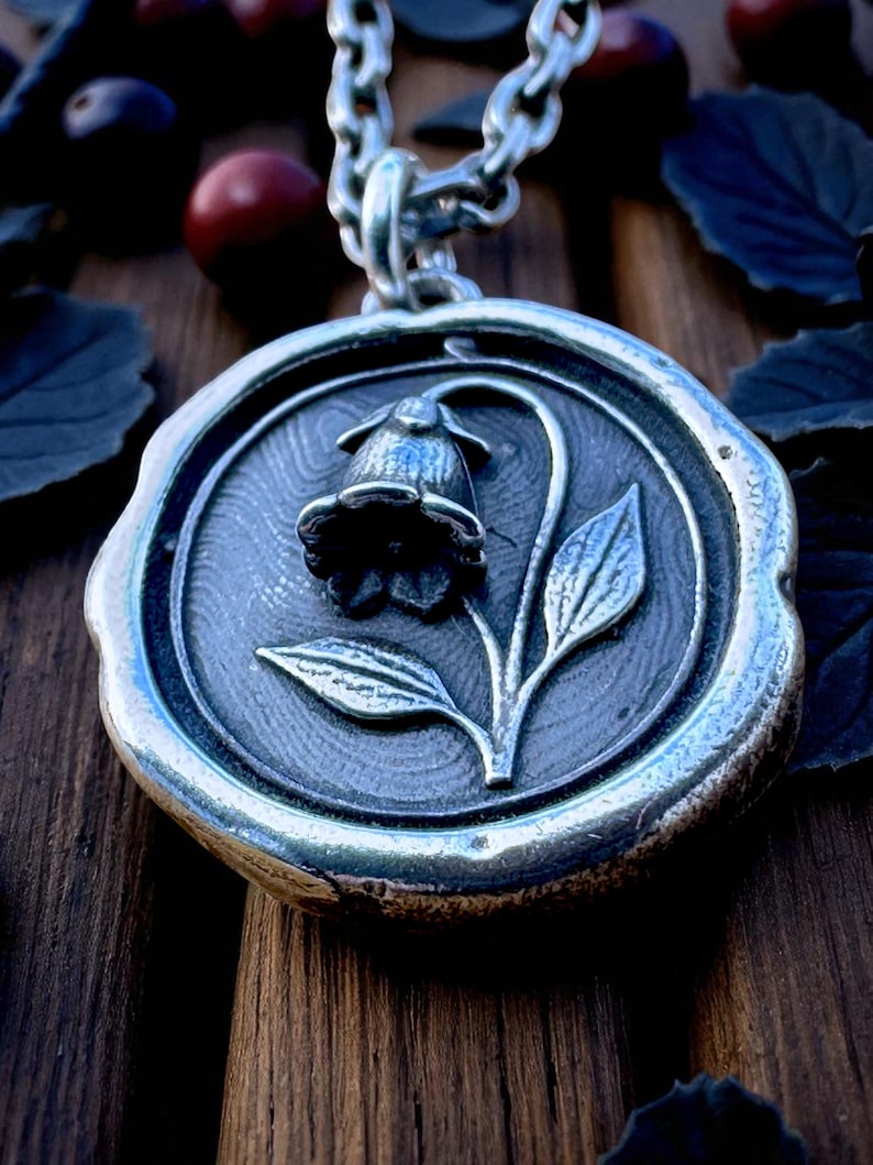 May include: A silver-toned pendant necklace featuring a floral design. The pendant has a circular shape with a raised border and a detailed image of a flower with leaves. The chain is also silver and has a link design.