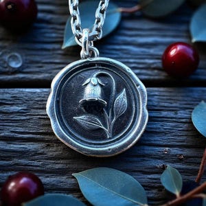 May include: A silver pendant necklace featuring a detailed floral design within a circular frame. The pendant hangs from a chain and is set against a backdrop of dark wood, red berries, and green leaves. The pendant's design includes a flower and leaves.