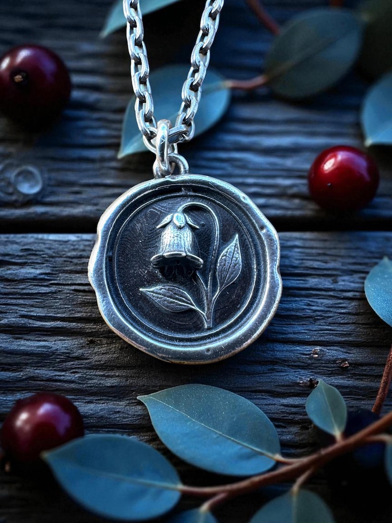 May include: A silver pendant necklace featuring a detailed floral design within a circular frame. The pendant hangs from a chain and is set against a backdrop of dark wood, red berries, and green leaves. The pendant's design includes a flower and leaves.