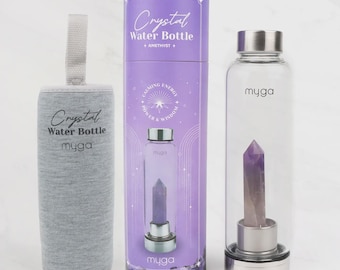 Puede incluir: Una botella de agua de cristal transparente con un cristal de amatista púrpura en el interior, junto a una funda de botella gris y una caja púrpura. La botella y la caja tienen el texto "Crystal Water Bottle" y la marca "myga".