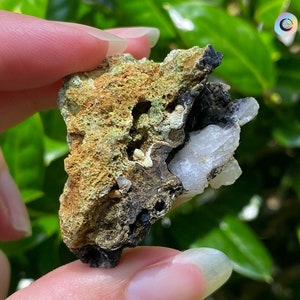 May include: A small, rough, green, black, and white rock with a crystalline texture. The rock is being held in a hand.