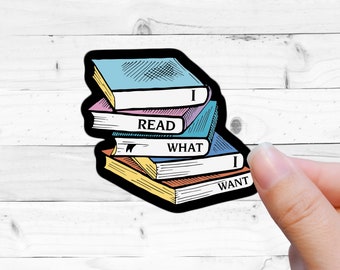 May include: A sticker featuring a stack of books with the words "I READ WHAT I WANT." The books have blue, pink, and yellow covers with black outlines. The sticker is held by a hand against a white wood background.