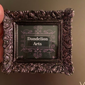 Könnte beinhalten: Ein kleiner schwarzer Bilderrahmen mit einem verzierten Design. Der Rahmen hat eine Glasfront und einen schwarzen Hintergrund mit einer weißen Aufschrift, die "Dandelion Arts" lautet.