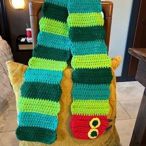 May include: A long, crocheted scarf featuring horizontal stripes in alternating shades of lime green, forest green, and teal. One end of the scarf is shaped like a red caterpillar head with two yellow and green circular eyes and small purple antennae.