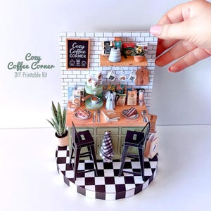 Może przedstawiać: Miniaturowy zestaw do druku DIY z kącikiem kawowym. Scena obejmuje ekspres do kawy, kubki, ciasta i tablicę z napisem "Cozy Coffee Corner". Zestaw znajduje się na czarno-białej szachownicy.