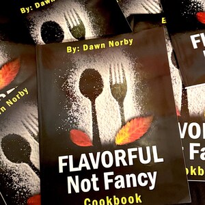以下が含まれることがあります： Dawn Norbyの「Flavorful Not Fancy Cookbook」というタイトルの料理本の束。黒い表紙にはスプーンとフォークのシルエットのデザイン、白い文字、秋の葉のアクセントが施されています。
