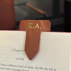 May include: A brown leather bookmark with the initials "K.A." in gold lettering is inserted into a book. The book has a pink cover and the pages are open to a chapter titled "Lottie".