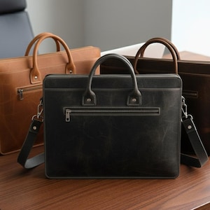 May include: Three leather briefcases in shades of brown and black sit on a wooden table. Each briefcase has a top handle, a front zipper pocket, and a detachable shoulder strap. The bags are designed for professional use.