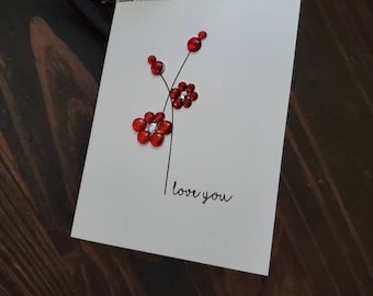 May include: A white greeting card with a floral design. The card features three red jeweled flowers with black stems and the handwritten phrase "love you". The card is on a dark brown wooden surface.