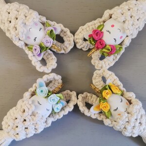 May include: Four handmade crocheted unicorn ornaments made with white yarn and colourful flower accents. Each ornament features a white egg-shaped base with painted eyes and a unicorn horn.