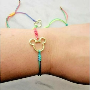 May include: A bracelet featuring a gold-toned metal charm in the shape of a mouse head silhouette. The charm is attached to a multi-colored cord made of thin, braided threads in shades of pink, green, and blue, with adjustable sliding knots.