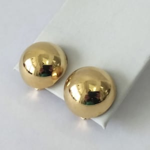 May include: A pair of polished gold-tone dome earrings with a smooth, reflective surface. Each earring features a rounded, hemispherical shape that catches the light, creating a bright, metallic sheen across the curved, solid gold-colored exterior.