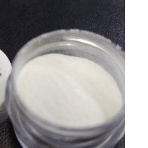 Op de afbeelding: Een klein, doorzichtig potje gevuld met wit poeder. Het potje heeft een schroefdop.