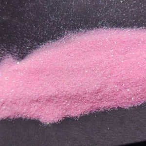 May include: A close-up of a pile of pink glitter. The glitter is a light, pastel pink color and has a sparkling, iridescent quality. The glitter is piled on a dark surface, creating a contrast.