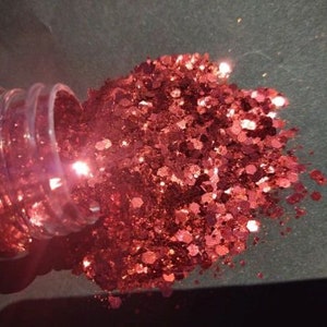 May include: A clear plastic bottle tipped over, spilling a large pile of sparkling red glitter. The glitter is in various shapes and sizes, reflecting light. The background is a dark, neutral color.