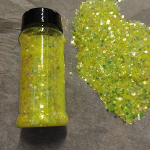 May include: A glass jar filled with yellow glitter with iridescent flecks. The glitter is also spilled out on a brown surface.