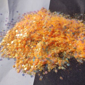May include: A pile of orange iridescent glitter with a holographic finish. The glitter is scattered on a white and black surface.