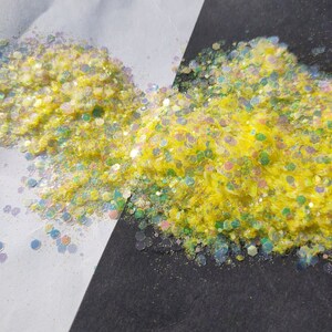May include: A close-up of yellow glitter with iridescent accents. The glitter is scattered on a white and black surface.