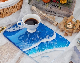 May include: A blue and white resin serving tray with a wave design. The tray is on a white lace tablecloth with a white cup of coffee and a small candle holder on the side.