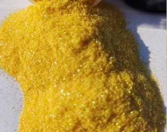 May include: Close-up of a pile of yellow glitter spilling from a clear container. The glitter is a bright, shimmering yellow with a fine texture. A black container is visible in the background.