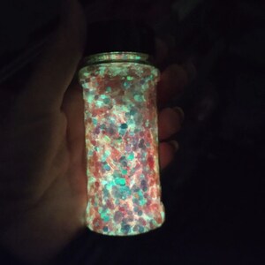 May include: A clear glass jar filled with colourful glitter that glows in the dark. The glitter is a mix of red, pink, blue, green, and white.