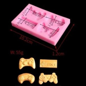 May include: A pink silicone mold for crafting, featuring four cavities in the shape of video game controllers and a game console. The mold measures 10.2 cm long, 1.2 cm thick, and weighs 55g. The cavities are labeled with measurements in centimeters.
