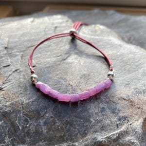 Könnte beinhalten: Ein Armband mit rosa Edelsteinen und einer weinroten Kordel. Das Armband besteht aus einer Reihe rechteckiger, rosa Edelsteine, die aneinandergereiht sind. Die Kordel ist mit einem Schiebeknoten verstellbar. Das Armband wird auf einer dunklen, strukturierten Oberfläche präsentiert.