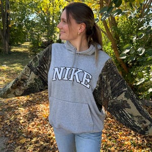 May include: A gray hooded sweatshirt featuring long sleeves with a brown and green forest camouflage pattern. The front of the garment displays the word NIKE in white block letters with a black outline, positioned above a front kangaroo pocket.