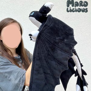 Può includere: Un peluche a forma di drago nero e bianco, con grandi ali strutturate. Il drago ha occhi blu e un ventre bianco. Il testo "Maro Licious" è nell'angolo in alto a destra. Il giocattolo viene tenuto in mano.