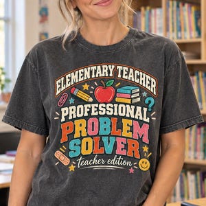 May include: A charcoal gray mineral-washed t-shirt featuring a colorful graphic with the text "ELEMENTARY TEACHER PROFESSIONAL PROBLEM SOLVER teacher edition" alongside illustrations of a red apple, a pencil, books, and a smiley face.