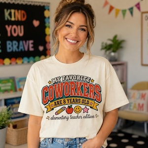 以下が含まれることがあります： クリーム色の半袖Tシャツ。赤と青の文字で「MY FAVORITE COWORKERS ARE 8 YEARS OLD」、その下に「elementary teacher edition」と書かれ、鉛筆、リンゴ、スマイリーフェイス、星のアイコンがデザインされています。