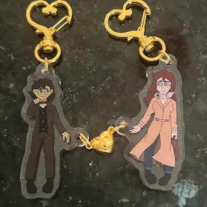 May include: Two matching keychains featuring cartoon illustrations of a man and a woman, connected by a gold heart charm. Each keychain has a gold heart-shaped clasp. The man wears a brown suit, and the woman wears a pink coat.
