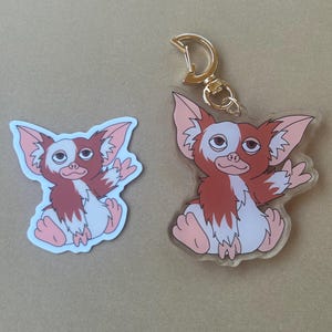May include: A sticker and a keychain featuring a cartoon gremlin character. The gremlin has brown and white fur, large ears, and is sitting with its arms outstretched. The keychain has a gold-colored clasp.