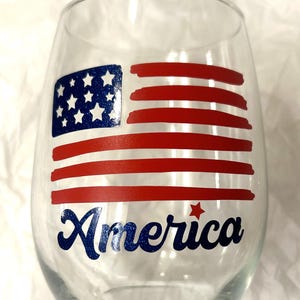 May include: Clear stemless wine glass with a patriotic design. The glass features a blue and white American flag and the word "America" in blue script. Red and white stripes complete the design.
