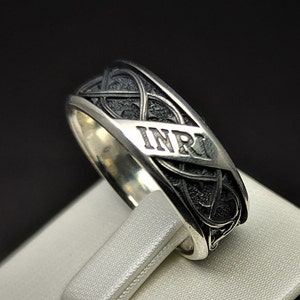May include: A silver ring featuring a black Celtic knot design with the inscription "INRI" on the band.