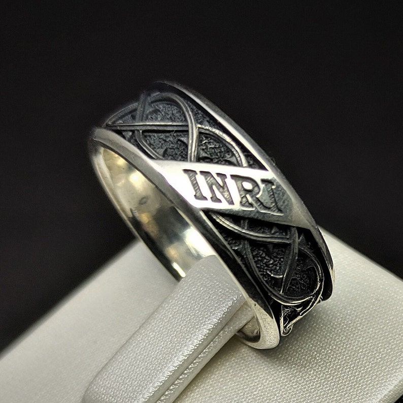 May include: A silver ring featuring a black Celtic knot design with the inscription "INRI" on the band.