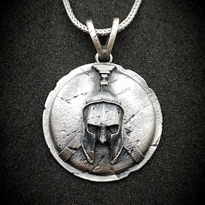 May include: Silver pendant necklace featuring a detailed, textured design of a Spartan helmet on a round shield.