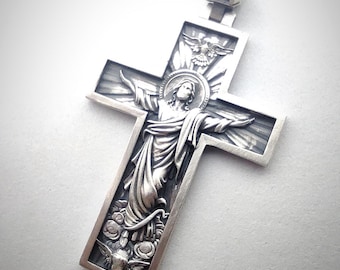 May include: A silver-colored pendant in the shape of a cross. The cross features a detailed relief of a religious figure with outstretched arms, surrounded by intricate designs. The bail of the pendant also has a cross design.
