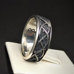May include: A silver ring featuring a black, intricate design. The inside of the band is engraved with an infinity symbol.
