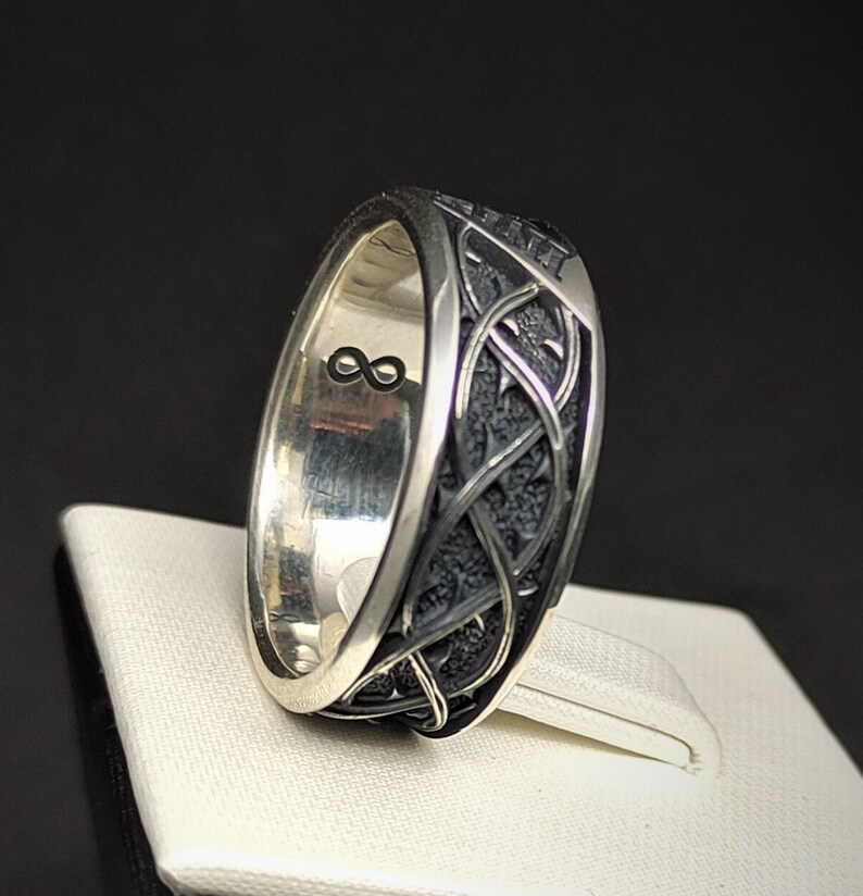 May include: A silver ring featuring a black, intricate design. The inside of the band is engraved with an infinity symbol.