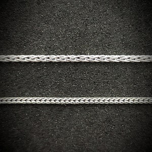 Może przedstawiać: Dwa srebrne łańcuszki z plecionym wzorem, leżące płasko na czarnej, fakturowanej powierzchni.