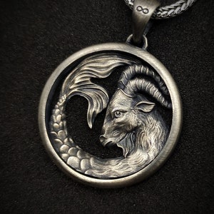 Może przedstawiać: Srebrny naszyjnik z zawieszką przedstawiającą szczeg&oacute;łowe przedstawienie znaku zodiaku Koziorożca. Zawieszka jest okrągła i przedstawia kozę z rybnym ogonem.