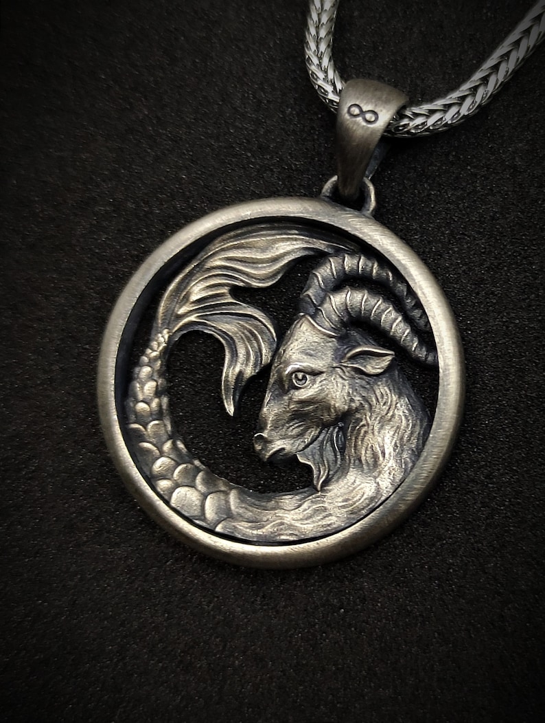Może przedstawiać: Srebrny naszyjnik z zawieszką przedstawiającą szczeg&oacute;łowe przedstawienie znaku zodiaku Koziorożca. Zawieszka jest okrągła i przedstawia kozę z rybnym ogonem.