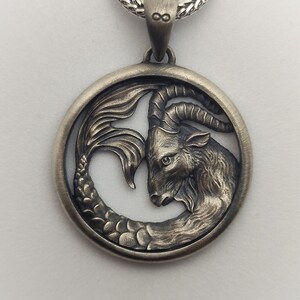 Może przedstawiać: Srebrny naszyjnik z zawieszką przedstawiającą szczeg&oacute;łowe przedstawienie znaku zodiaku Koziorożca, kozy z rybnym ogonem, w okrągłej ramie.