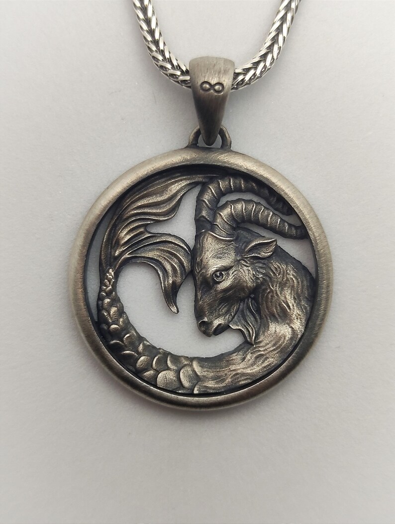 Może przedstawiać: Srebrny naszyjnik z zawieszką przedstawiającą szczeg&oacute;łowe przedstawienie znaku zodiaku Koziorożca, kozy z rybnym ogonem, w okrągłej ramie.