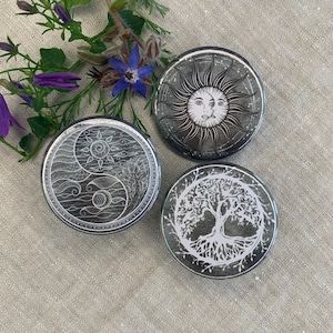 Könnte beinhalten: Drei runde schwarz-weiße Anstecknadeln mit verschiedenen Designs. Der erste Knopf zeigt ein Yin-Yang-Symbol mit einem Sonnen- und Monddesign. Der zweite Knopf zeigt ein Lebensbaumdesign. Der dritte Knopf zeigt eine Sonne mit einem Gesicht.
