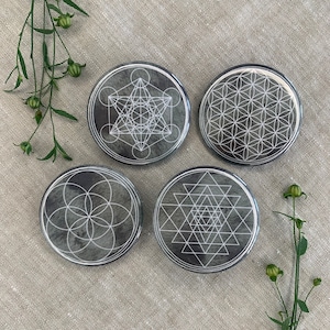 Op de afbeelding: Vier ronde onderzetters met een grijze achtergrond en witte geometrische ontwerpen. De ontwerpen omvatten een bloem des levens, een heilig geometrisch patroon, een Sri Yantra en een zaad van het leven.