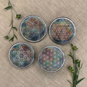 Op de afbeelding: Vier iriserende onderzetters met verschillende geometrische ontwerpen. Elke onderzetter heeft een wit geometrisch patroon op een donkere, iriserende achtergrond.
